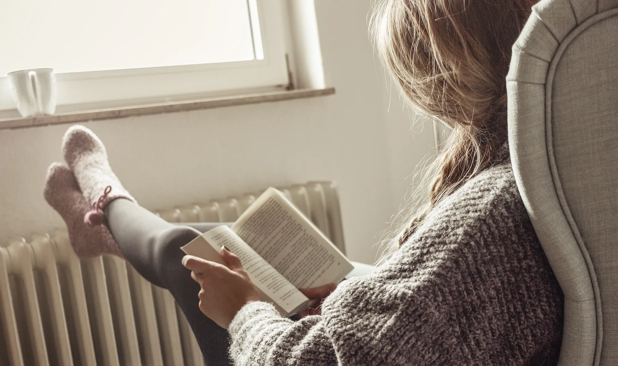 Eine Person in einem kuscheligen Pullover und Socken sitzt gemütlich in einem Sessel und liest ein Buch am Fenster. Sanftes Licht, eine entspannte Atmosphäre und eine Tasse in der Nähe vermitteln Gemütlichkeit.